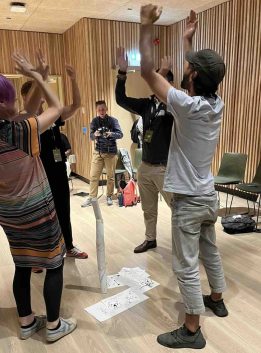 Four people reach their arms to the sky as they stand around a paper tower sculpture