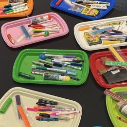 Brightly colored trays placed on a black tablecloth contain a range of art materials